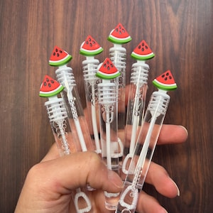 May include: A collection of clear bubble wands, each topped with a watermelon slice design. The wands are held in a hand against a dark wood background. The watermelon slices feature red, green, and black details.