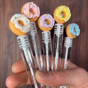 May include: A collection of bubble wands topped with miniature, colorful donut designs. Each donut features a different frosting color, including pink, yellow, purple, and blue, with sprinkles. The wands are clear plastic with a bubble solution.