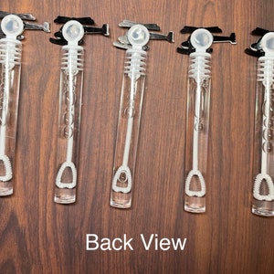 May include: Five clear plastic bubble wands with white caps and internal wands, each topped with a black helicopter-shaped handle. The wands are filled with bubble solution and feature a ribbed design on the internal wand. The text Back View is visible.