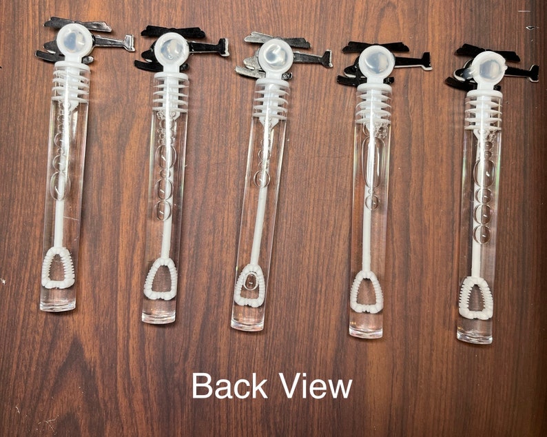 May include: Five clear plastic bubble wands with white caps and internal wands, each topped with a black helicopter-shaped handle. The wands are filled with bubble solution and feature a ribbed design on the internal wand. The text Back View is visible.