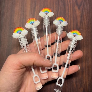 May include: Six clear bubble wands with rainbow and cloud toppers. Each wand has a white handle and a bubble loop. The rainbow toppers feature vibrant colors, and the wands are held against a dark wood background.