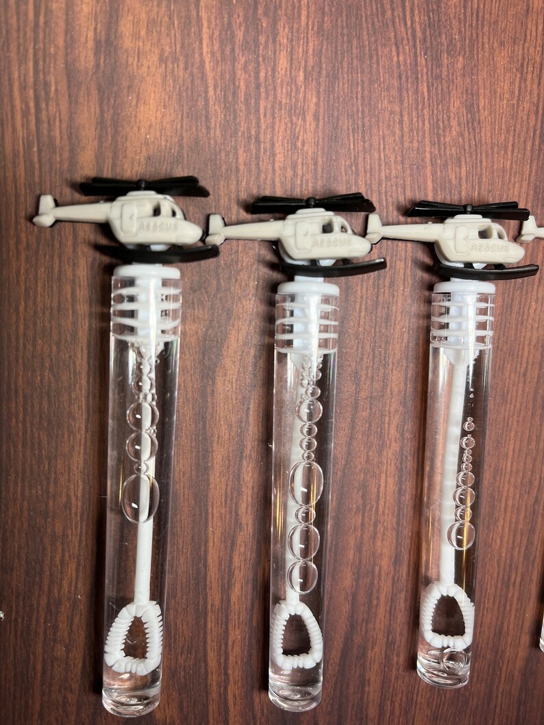 May include: Three clear plastic bubble wands, each topped with a light gray helicopter featuring black rotors and the word RESCUE embossed on the side. The wands contain clear bubble solution and a white internal wand with a circular loop at the base.
