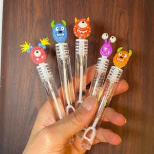 May include: Five bubble wands with colorful monster-themed toppers. Each wand has a clear plastic tube with a white handle and a bubble-making loop. The monster heads are various colors, including blue, orange, and purple.