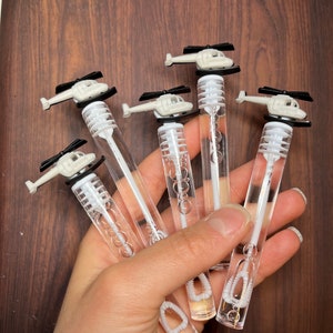 May include: Five clear plastic bubble wands, each topped with a small cream-colored helicopter featuring black rotor blades. The wands are filled with clear bubble solution and contain a white plastic wand applicator inside the cylindrical tube.