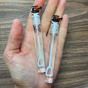 May include: Two clear plastic bubble wands featuring a small brown and black helicopter topper. Each wand has a white internal stem with a circular loop at the bottom for blowing bubbles, topped with a detailed miniature helicopter figurine.