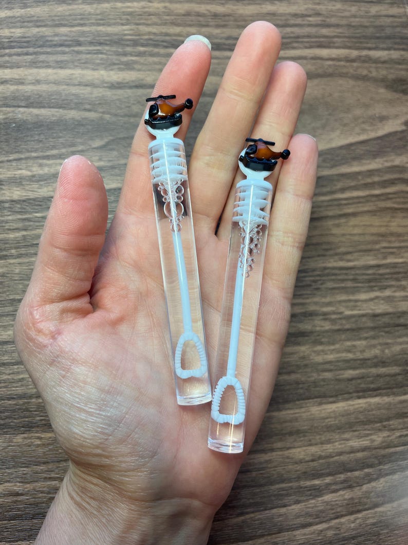 May include: Two clear plastic bubble wands featuring a small brown and black helicopter topper. Each wand has a white internal stem with a circular loop at the bottom for blowing bubbles, topped with a detailed miniature helicopter figurine.