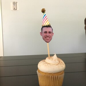 Puede incluir: Un cupcake blanco con una cobertura de az&uacute;car moreno y un remolino de glaseado blanco. Un gorro de fiesta con un patr&oacute;n de rayas arco&iacute;ris y un pomp&oacute;n dorado est&aacute; en la parte superior del cupcake. La cara de una persona est&aacute; en la parte delantera del gorro.