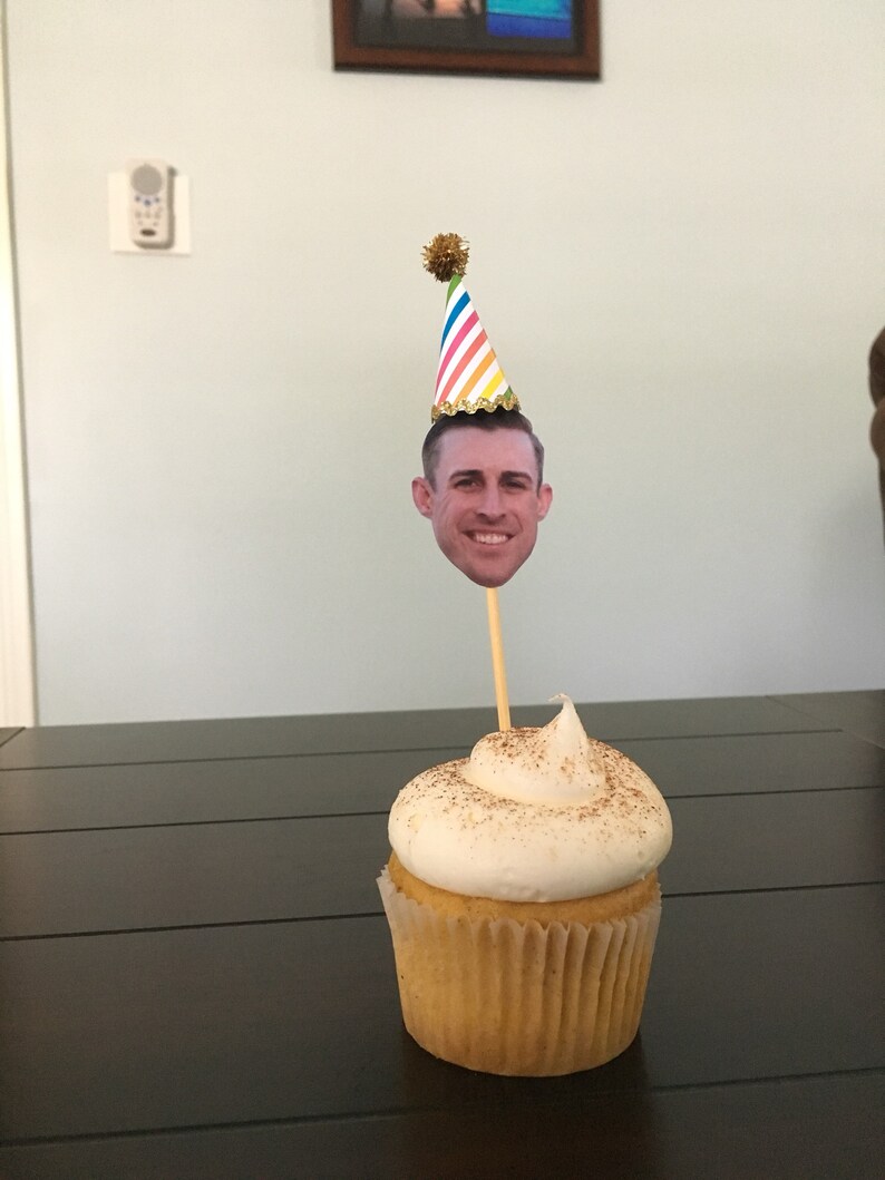 Puede incluir: Un cupcake blanco con una cobertura de az&uacute;car moreno y un remolino de glaseado blanco. Un gorro de fiesta con un patr&oacute;n de rayas arco&iacute;ris y un pomp&oacute;n dorado est&aacute; en la parte superior del cupcake. La cara de una persona est&aacute; en la parte delantera del gorro.