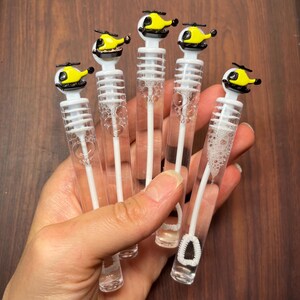 May include: Five clear plastic bubble wands, each topped with a small yellow and black helicopter figurine. The wands contain clear bubble solution and feature a white internal wand with a circular loop at the bottom for blowing bubbles.