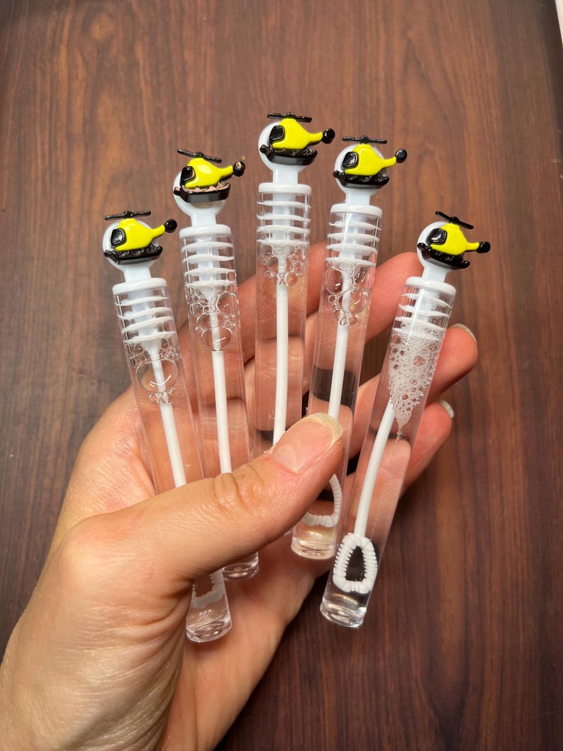 May include: Five clear plastic bubble wands, each topped with a small yellow and black helicopter figurine. The wands contain clear bubble solution and feature a white internal wand with a circular loop at the bottom for blowing bubbles.