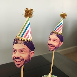 Puede incluir: Dos cupcakes con glaseado blanco y chispas de colores cubiertos con sombreros de fiesta. Los sombreros est&aacute;n decorados con la cara de una persona y un borde de purpurina dorada.
