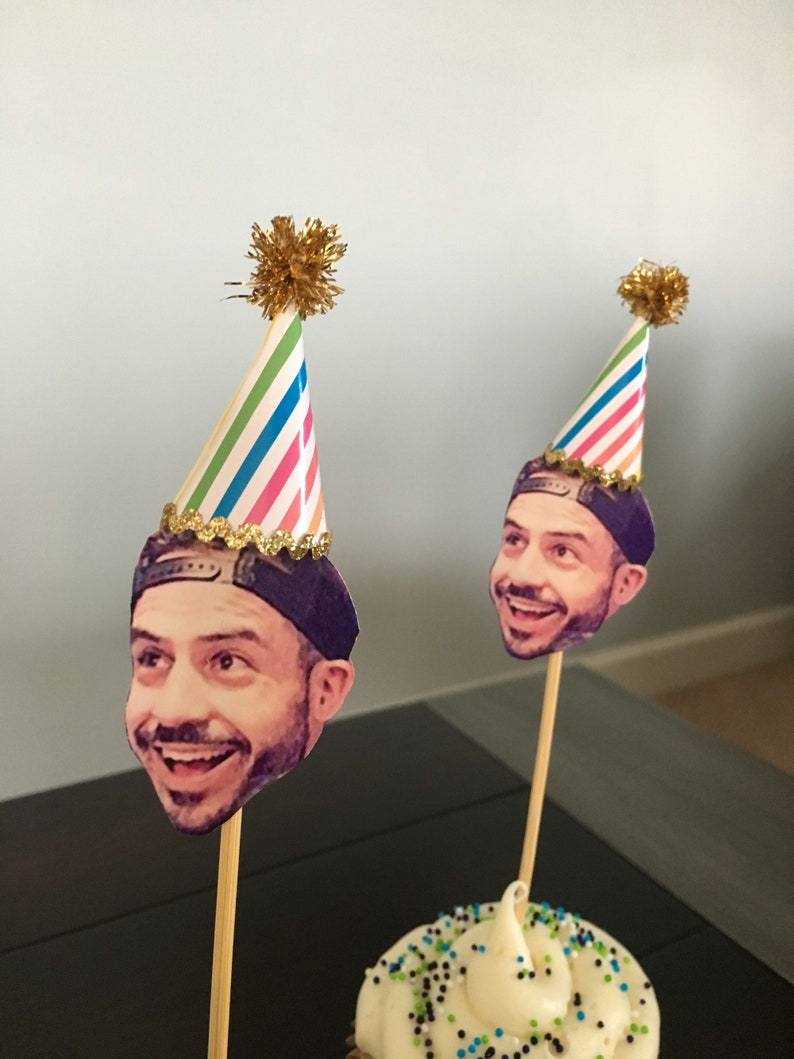 Puede incluir: Dos cupcakes con glaseado blanco y chispas de colores cubiertos con sombreros de fiesta. Los sombreros est&aacute;n decorados con la cara de una persona y un borde de purpurina dorada.