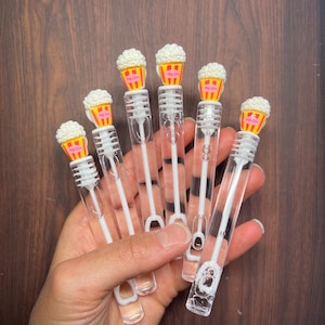 May include: Five clear plastic bubble wands featuring a miniature popcorn bucket topper. Each topper is white and yellow with red stripes and the text Pop Corn. The wands have a white internal stem and a circular loop at the bottom for blowing bubbles.