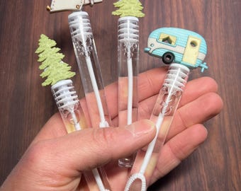 May include: Four clear plastic bubble wands with different themed toppers. The toppers are a green pine tree, a white tent, a green pine tree, and a blue and white camper.