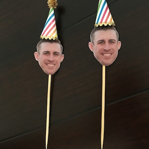 Puede incluir: Dos adornos para pasteles con la cara de una persona y un gorro de fiesta colorido en la parte superior de un palo de madera.