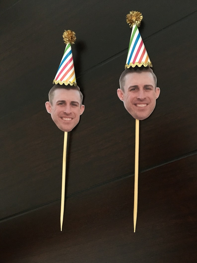 Puede incluir: Dos adornos para pasteles con la cara de una persona y un gorro de fiesta colorido en la parte superior de un palo de madera.