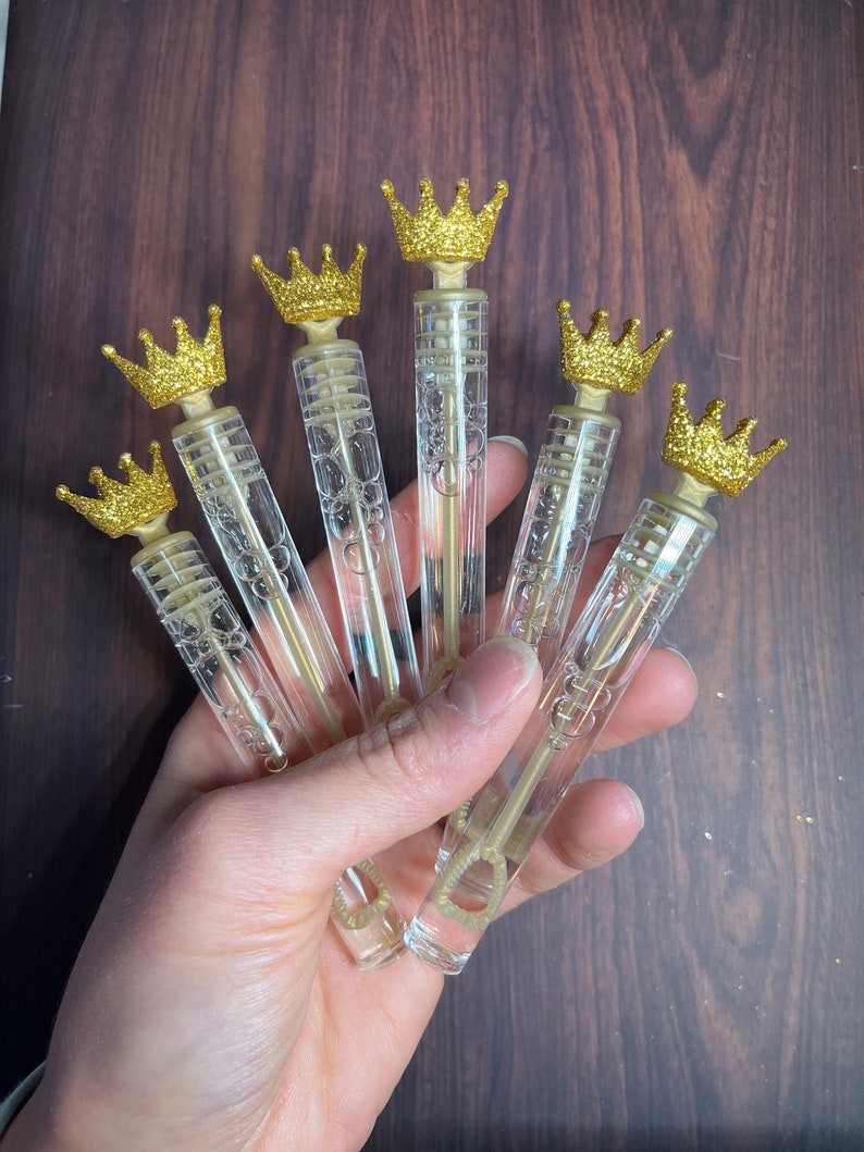 May include: Six clear plastic bubble wands with gold glitter crowns on top. The wands are held in a hand.