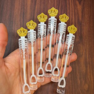 May include: Six clear plastic bubble wands with gold glitter crown toppers. The wands have a white plastic loop at the end for blowing bubbles.