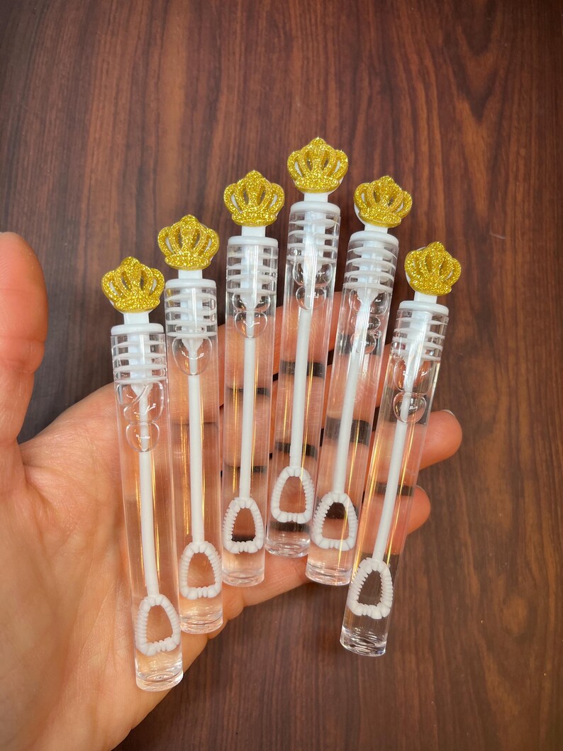 May include: Six clear plastic bubble wands with gold glitter crown toppers. The wands have a white plastic loop at the end for blowing bubbles.