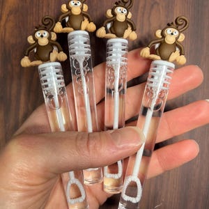 May include: Four clear bubble wands with monkey toppers. Each wand has a clear plastic tube with a white handle and a bubble loop. The monkey toppers are brown and tan, with detailed facial features and curly tails.