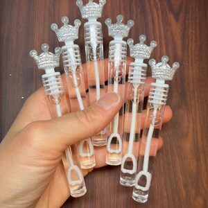 May include: Six clear plastic bubble wands with silver glitter crowns on top. The wands are held in a hand.