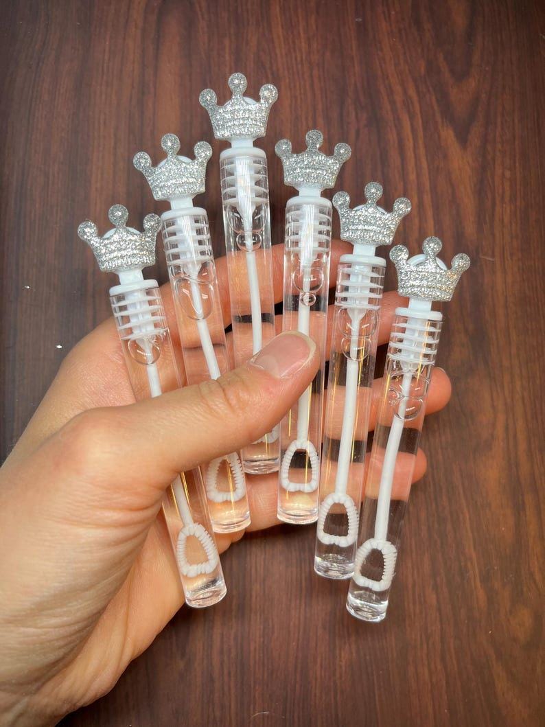 May include: Six clear plastic bubble wands with silver glitter crowns on top. The wands are held in a hand.
