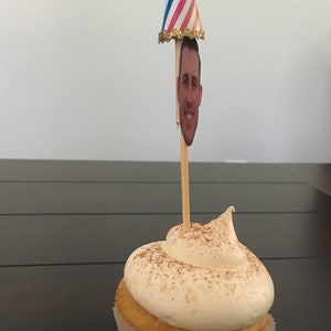 Puede incluir: Un cupcake de vainilla con glaseado blanco y una pizca de canela en la parte superior. Un gorro de fiesta con un recorte de cara est&aacute; en un palo en el glaseado.