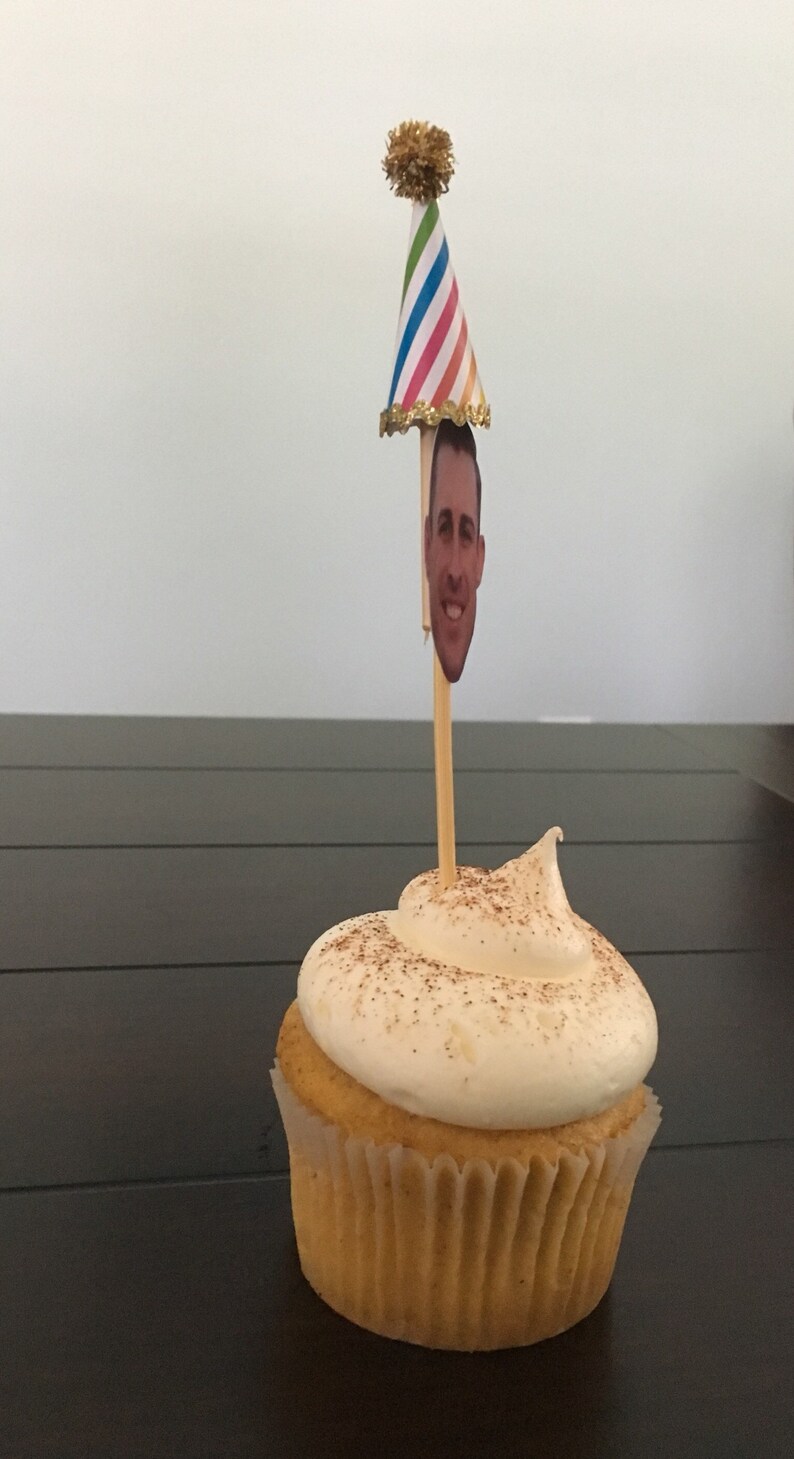 Puede incluir: Un cupcake de vainilla con glaseado blanco y una pizca de canela en la parte superior. Un gorro de fiesta con un recorte de cara est&aacute; en un palo en el glaseado.