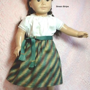 May include: A doll wearing a white blouse with lace trim, a green striped skirt with a green sash, and brown sandals. The doll has brown hair and brown eyes. The text "Green Stripe" is visible in the image.