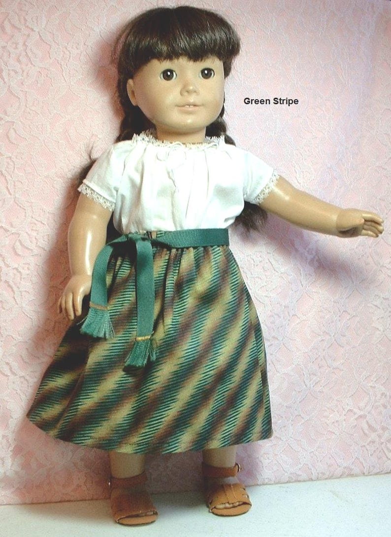 May include: A doll wearing a white blouse with lace trim, a green striped skirt with a green sash, and brown sandals. The doll has brown hair and brown eyes. The text "Green Stripe" is visible in the image.