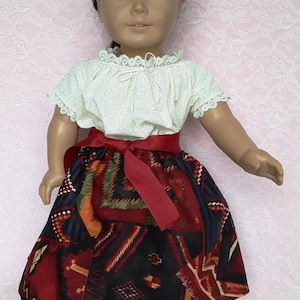 May include: A doll wearing a white blouse with lace trim, a red ribbon belt, and a colourful skirt with a geometric pattern. The doll is wearing brown moccasins with turquoise beads. The doll has brown hair and brown eyes. The text "Navajo Blanket" is visible in the image.