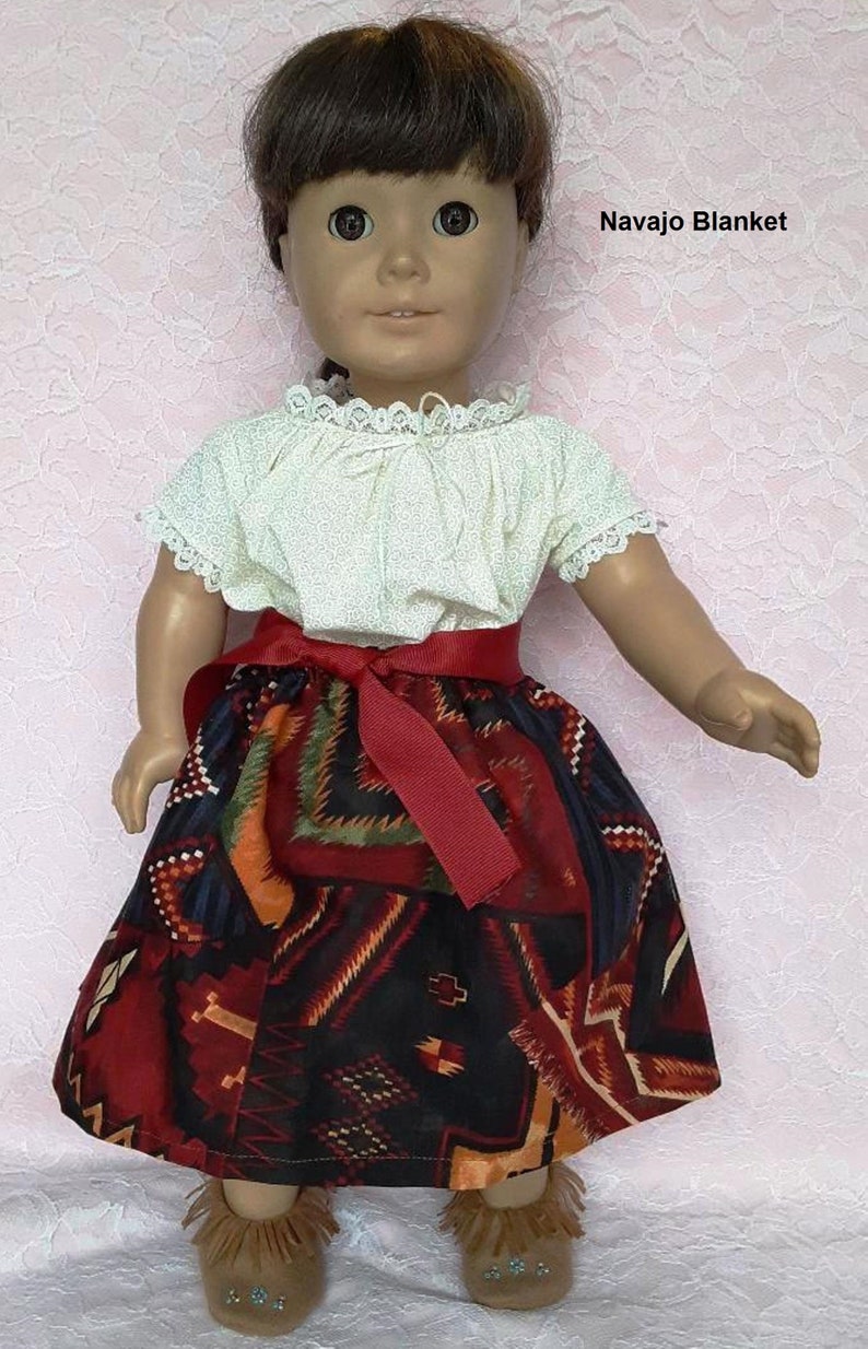 May include: A doll wearing a white blouse with lace trim, a red ribbon belt, and a colourful skirt with a geometric pattern. The doll is wearing brown moccasins with turquoise beads. The doll has brown hair and brown eyes. The text "Navajo Blanket" is visible in the image.