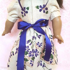May include: A white dress with a purple floral pattern and a blue ribbon tied around the waist. The dress has short sleeves and a gathered skirt.