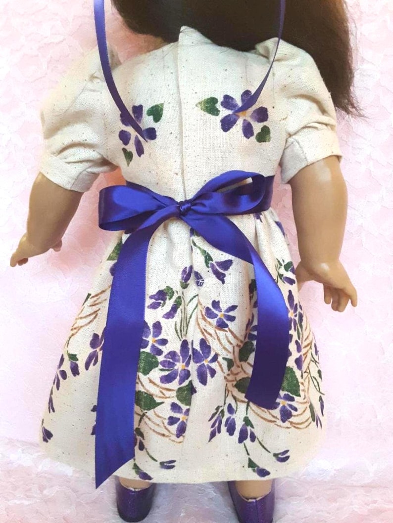 May include: A white dress with a purple floral pattern and a blue ribbon tied around the waist. The dress has short sleeves and a gathered skirt.