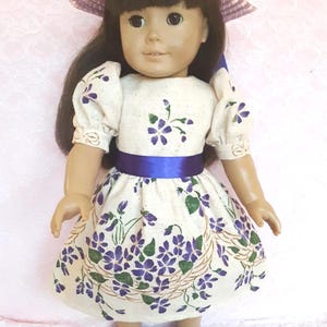 May include: A doll wearing a white dress with purple flowers and a blue sash. The doll is also wearing a purple hat and purple shoes.