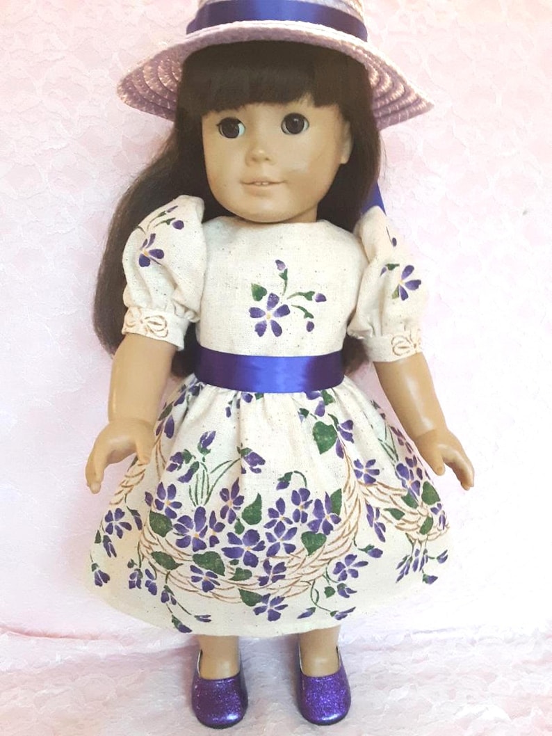 May include: A doll wearing a white dress with purple flowers and a blue sash. The doll is also wearing a purple hat and purple shoes.