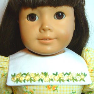 May include: A doll wearing a yellow and white gingham dress with a white collar and embroidered floral trim. The doll has brown hair and brown eyes.