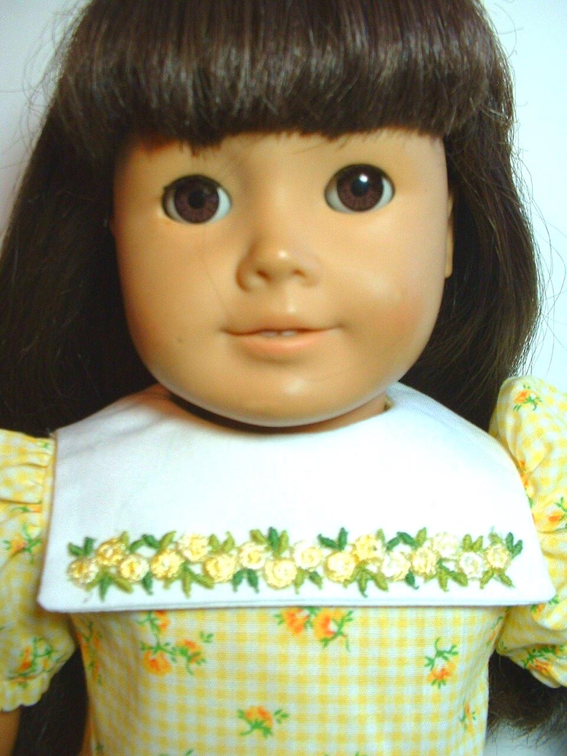 May include: A doll wearing a yellow and white gingham dress with a white collar and embroidered floral trim. The doll has brown hair and brown eyes.