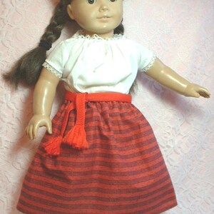 May include: A doll wearing a white blouse with a red striped skirt and a red belt with a tassel. The doll has brown hair in braids and brown eyes. The text "Red Stripe" is visible in the upper right corner of the image.