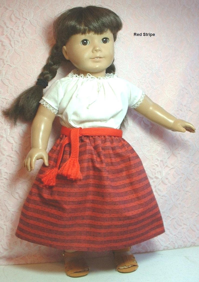 May include: A doll wearing a white blouse with a red striped skirt and a red belt with a tassel. The doll has brown hair in braids and brown eyes. The text "Red Stripe" is visible in the upper right corner of the image.