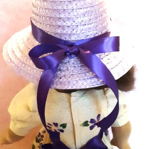 May include: A lavender straw hat with a wide brim and a purple satin ribbon tied in a bow at the back. The hat is worn by a doll with a yellow dress with purple flowers.