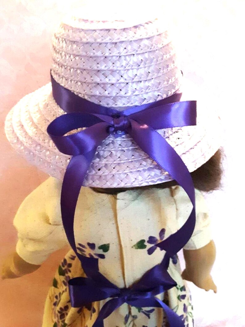 May include: A lavender straw hat with a wide brim and a purple satin ribbon tied in a bow at the back. The hat is worn by a doll with a yellow dress with purple flowers.