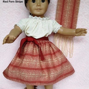 May include: A doll wearing a red and cream striped skirt with a red belt, a white blouse with lace trim, and brown moccasins. The doll is holding a red and cream striped scarf.