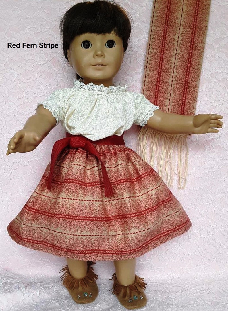 May include: A doll wearing a red and cream striped skirt with a red belt, a white blouse with lace trim, and brown moccasins. The doll is holding a red and cream striped scarf.