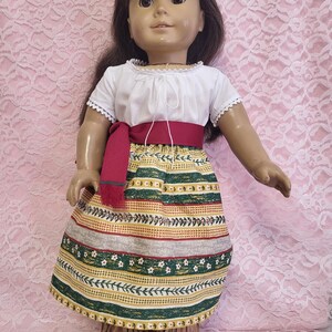 May include: A doll wearing a white peasant top with a red sash and a green and gold patterned skirt. The skirt has a floral design and vines. The doll has brown hair and brown eyes. Green/Gold Vines.