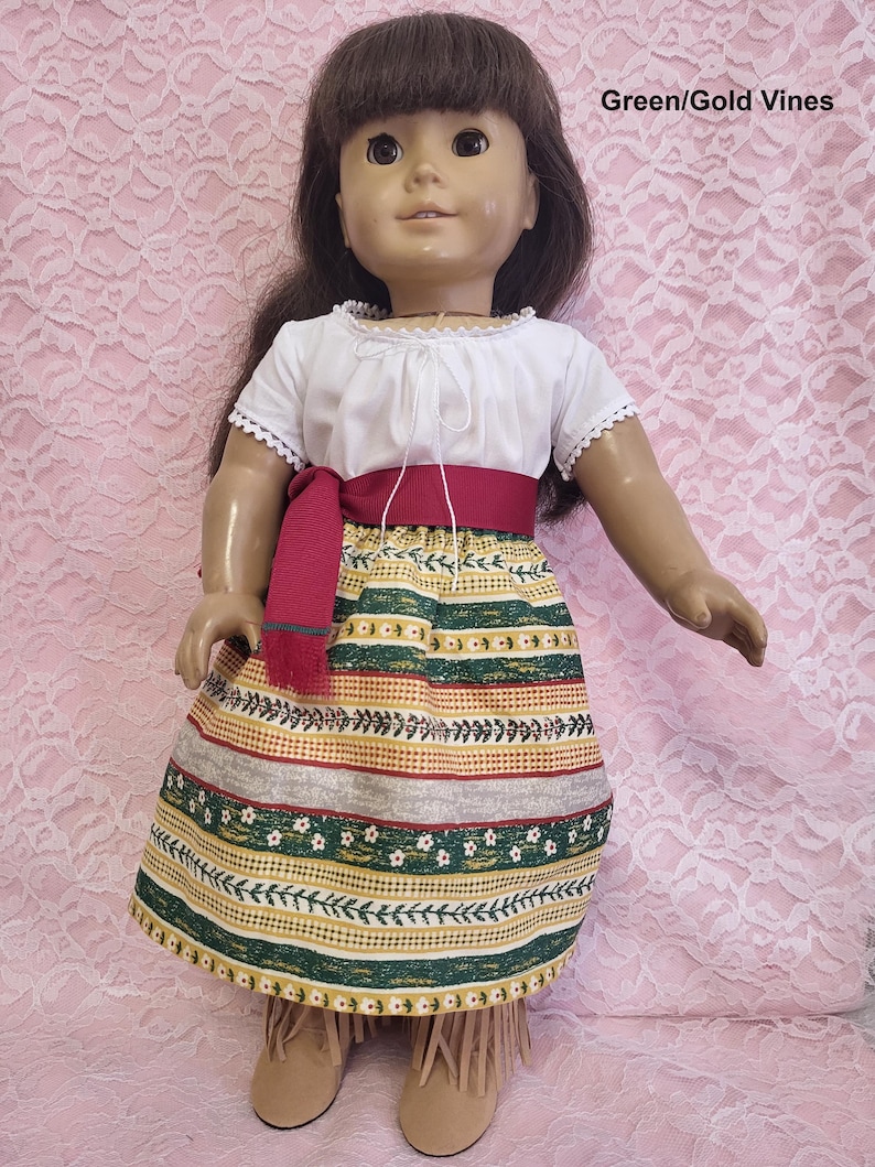 May include: A doll wearing a white peasant top with a red sash and a green and gold patterned skirt. The skirt has a floral design and vines. The doll has brown hair and brown eyes. Green/Gold Vines.