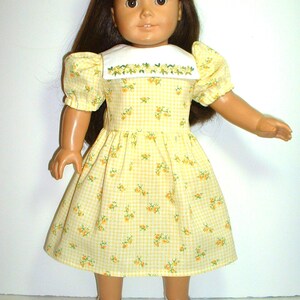 May include: A yellow and white gingham dress with a floral print and a white embroidered collar. The dress is worn by a doll with brown hair and brown eyes.
