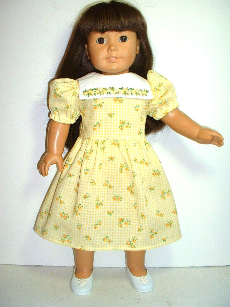 May include: A yellow and white gingham dress with a floral print and a white embroidered collar. The dress is worn by a doll with brown hair and brown eyes.