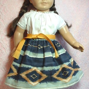 May include: A doll wearing a white blouse with a lace trim, a blue and orange ikat skirt, a yellow belt, and brown sandals. The doll has brown hair in braids and brown eyes. The background is a pink and white floral pattern.
