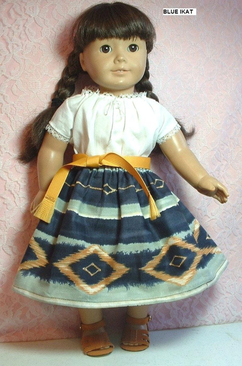 May include: A doll wearing a white blouse with a lace trim, a blue and orange ikat skirt, a yellow belt, and brown sandals. The doll has brown hair in braids and brown eyes. The background is a pink and white floral pattern.